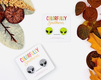 May include: Two pairs of stud earrings on white display cards labeled Colorfully Southern. One pair features bright green alien faces with black eyes, and the other pair features silver glitter alien faces with black eyes. Each card says Handmade.