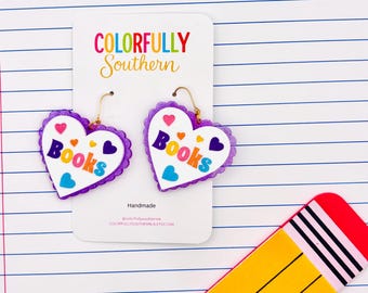 May include: A pair of heart-shaped earrings with a white center, purple scalloped edges, and gold-toned hooks. The earrings feature the word "Books" in multicolored letters, surrounded by small red, orange, and blue heart icons on a display card.