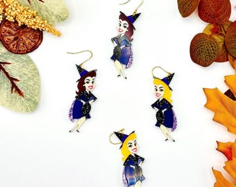 May include: Four dangle earrings featuring cartoon witch figures with gold-tone hooks. Two figures have brown hair and two have blonde hair. Each witch wears a dark blue glittery dress, a matching pointed hat, and a purple cape with a glittery finish.
