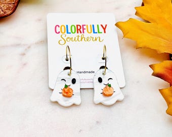 May include: A pair of white ghost-shaped earrings with a marbled texture, each featuring a small orange pumpkin graphic and a winking face. The earrings hang from gold-toned hoops on a white display card labeled Colorfully Southern.