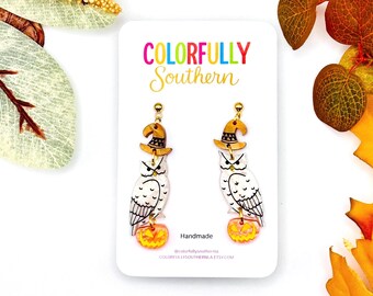 May include: A pair of dangle earrings featuring white owls with black line details, each wearing a tan witch hat and perched atop a small orange jack-o'-lantern. The earrings are displayed on a white card labeled Colorfully Southern.