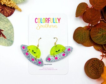 May include: A pair of dangle earrings shaped like silver glitter UFOs with neon pink dots and a bright green translucent dome featuring a black alien face. The earrings are attached to a white display card with the text COLORFULLY Southern.