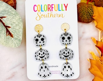 May include: Dangle earrings featuring a gold glitter stud and three stacked silver glitter charms in the shapes of a sugar skull, a jack-o'-lantern, and a ghost, all with black printed details, displayed on a white card labeled Colorfully Southern.