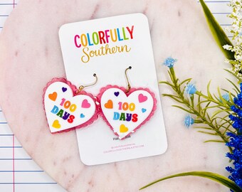 May include: A pair of heart-shaped dangle earrings featuring a white surface with the text 100 DAYS in colorful letters, surrounded by small multi-colored hearts. The earrings have a pink scalloped border and gold-toned metal hooks.