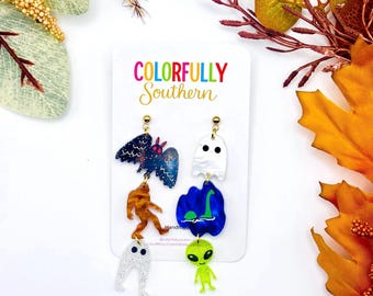 May include: A pair of mismatched dangle earrings on a white display card labeled Colorfully Southern. One earring features a dark blue bat, a brown Bigfoot, and a white glittery ghost. The other features a white ghost, a blue Loch Ness monster, and a green alien.