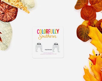 May include: A pair of small, white, glittery ghost-shaped stud earrings displayed on a square white card. The card features the text Colorfully Southern in colorful letters, the word Handmade, and a social media handle in small black print.
