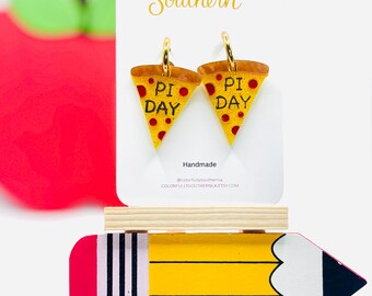 May include: A pair of gold-tone hoop earrings shaped like pizza slices with yellow crusts, red pepperoni dots, and the text PI DAY printed in black. The earrings are displayed on a white card labeled COLORFULLY SOUTHERN above a wooden pencil prop.