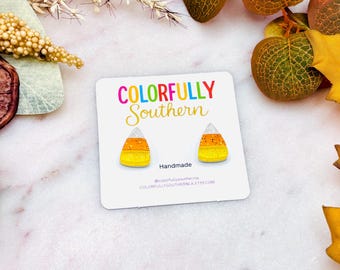 May include: A pair of candy corn shaped stud earrings on a white display card with the text COLORFULLY SOUTHERN. The earrings feature a white top, orange middle, and yellow bottom with a glittery finish. The card also includes the text Handmade.