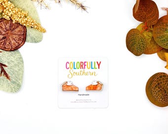 May include: A pair of orange pumpkin pie slice earrings with white whipped cream toppings, displayed on a white card labeled Colorfully Southern. The earrings feature a marbled orange texture and are positioned side by side on the branded card.