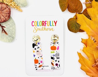 May include: A pair of rectangular dangle earrings featuring a white background with colorful Halloween-themed patterns including ghosts, pumpkins, skulls, and spiders. The earrings are attached to a white display card labeled COLORFULLY SOUTHERN.