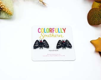 May include: A pair of black glitter bat stud earrings with red eyes displayed on a white card featuring the text Colorfully Southern Handmade and the website address. The earrings are shaped like small bats with detailed wing patterns and pointed ears.