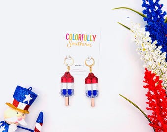 May include: A pair of dangle earrings shaped like red, white, and blue glittery popsicles with gold-toned hoops. The earrings are attached to a white display card featuring the text Colorfully Southern and Handmade in black and gold lettering.