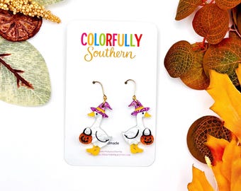 May include: A pair of white goose earrings with yellow beaks and feet, each wearing a purple glitter witch hat and holding a small orange jack-o'-lantern bucket. The earrings are displayed on a white card labeled Colorfully Southern.