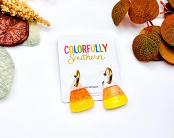 May include: A pair of candy corn shaped earrings featuring white, orange, and yellow horizontal stripes with a glitter finish. The earrings are attached to a white display card with the text Colorfully Southern printed in colorful letters.