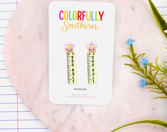 May include: A pair of dangle earrings on a white display card featuring the text COLORFULLY SOUTHERN. Each earring has a light purple flower stud attached to a yellow rectangular charm shaped like a ruler with the black text YOU RULE.