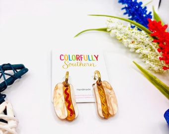 May include: A pair of dangle earrings shaped like hot dogs with cream-colored buns and gold-toned hoops. One earring features a red wavy line, and the other has a yellow wavy line. They are attached to a white display card labeled Colorfully Southern.