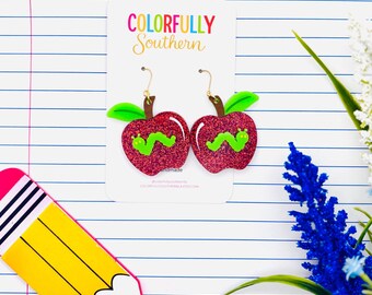 May include: A pair of dangle earrings featuring red glitter apples with green leaves and a small green caterpillar on each. The earrings are attached to a white display card with the text COLORFULLY SOUTHERN printed in colorful letters.