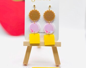 May include: Dangle earrings on a white display card labeled Colorfully Southern. Each earring features a tan scalloped cookie charm, a light pink circular bubble charm, and a bright yellow square charm. A yellow pencil graphic is below.