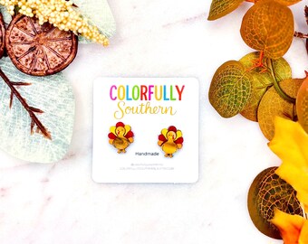 May include: A pair of small turkey-shaped stud earrings displayed on a white card featuring the text Colorfully Southern and Handmade. The earrings are yellow, red, and orange with a glossy finish, positioned centrally on the branded white card.