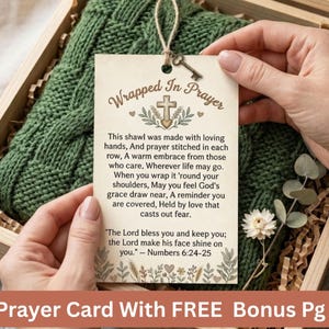 Op de afbeelding: Een groene gebreide sjaal in een houten doos met een label met de tekst "Wrapped in Prayer". Het label heeft een kruis- en bloemenontwerp en een sleutelbedel. De tekst op het label bevat een bijbelvers uit Numeri 6:24-25.