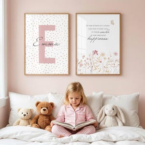 Könnte beinhalten: Zwei gerahmte Wandbilder. Das linke Bild zeigt einen großen rosa Buchstaben E mit dem Namen Emma in Schreibschrift und goldenen Punkten. Das rechte Bild zeigt den Text In the hands of a child lies the future and in their smile the greatest happiness mit Blumenillustrationen.