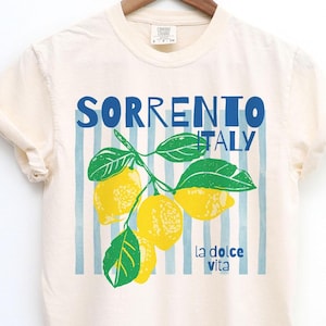 Puede incluir: Camiseta color crema con las palabras "SORRENTO ITALY" en letras azules. Debajo, un gr&aacute;fico de limones y hojas, con "la dolce vita" impreso debajo. La camiseta est&aacute; colgada en una percha de madera. El dise&ntilde;o tiene rayas verticales azules.