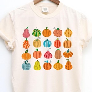 May include: A cream-colored short-sleeve t-shirt featuring a grid of twenty illustrated pumpkins in various colors, shapes, and patterns, including orange, blue, pink, yellow, and green, arranged in five rows and four columns on the front.