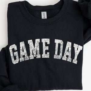 May include: Black sweatshirt with the text "GAME DAY" in distressed white lettering. The crew neck sweatshirt is folded, with the sleeves neatly tucked in. The Gildan brand tag is visible at the neckline. A casual, comfortable apparel item.