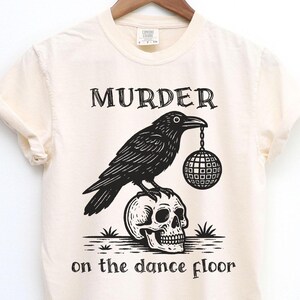 May include: Cream-colored t-shirt featuring a black graphic of a raven perched atop a skull, holding a disco ball. The text "MURDER" is above the raven, and "on the dance floor" is below. The shirt is hanging on a wooden hanger.