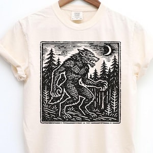 Puede incluir: Camiseta color crema con una ilustraci&oacute;n en blanco y negro de un hombre lobo en un bosque bajo una luna creciente. El dise&ntilde;o est&aacute; en un marco cuadrado, con un estilo de grabado en madera vintage. La camiseta est&aacute; colgada en una percha de madera.