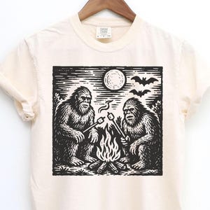 May include: Cream-colored t-shirt featuring a black and white graphic of two Bigfoot creatures roasting marshmallows over a campfire under a full moon. Bats are silhouetted in the sky above the moon. The shirt is hanging on a wooden hanger.