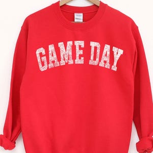 May include: A bright red sweatshirt with the text "GAME DAY" in distressed white lettering. The crew neck sweatshirt is hanging on a wooden hanger. The sleeves are cuffed at the wrists.