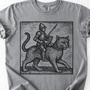 Puede incluir: Camiseta gris con una ilustraci&oacute;n en blanco y negro de un caballero montando un gato grande, sosteniendo una lanza y un escudo. El gr&aacute;fico est&aacute; enmarcado en un cuadrado. La camiseta es de material suave.