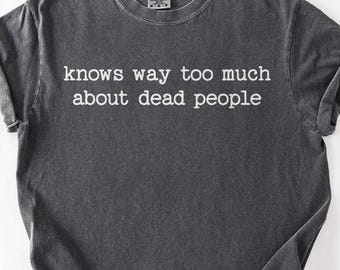 Pode incluir: T-shirt cinzenta escura com o texto branco "knows way too much about dead people". A camisa tem gola redonda e mangas enroladas. O tecido tem um aspeto vintage ligeiramente desbotado.