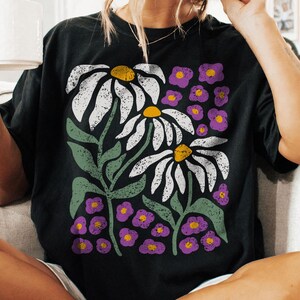 May include: Black t-shirt featuring a floral design with white daisies, yellow centers, and purple accent flowers. The graphic tee has green stems and leaves. The person is holding a white mug.