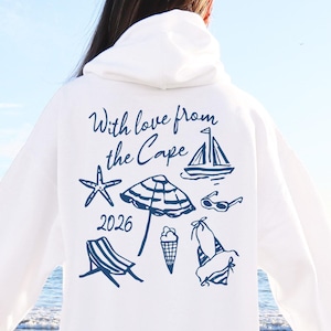 May include: White hooded sweatshirt with a beach-themed graphic in navy blue. The design includes a sailboat, starfish, beach umbrella, sunglasses, beach chair, ice cream cone, and a swimsuit. The text "With love from the Cape 2026" is also included.