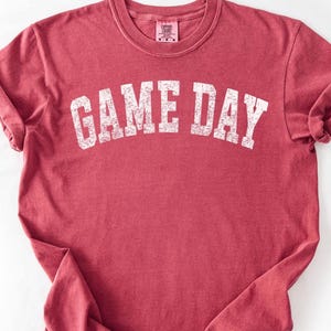 May include: A maroon t-shirt featuring the white text "GAME DAY" in an arched design. The shirt has a crew neck and rolled-up sleeves. The fabric has a slightly faded appearance, suggesting a vintage style.