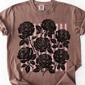 May include: A brown t-shirt featuring a black rose graphic with pink vertical stripes. The roses are detailed with leaves and stems. The shirt has a crew neck and short sleeves.
