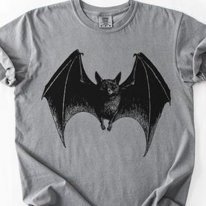 May include: A gray t-shirt featuring a detailed black illustration of a bat with outstretched wings. The bat is centered on the shirt, showcasing its wingspan. The shirt has a classic crew neck and short sleeves.