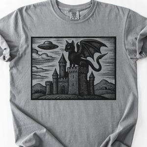 May include: Gray t-shirt featuring a black and white graphic of a winged cat perched atop a castle, with a UFO in the sky. The design has a vintage, woodcut style. The shirt is made of a soft, comfortable material.