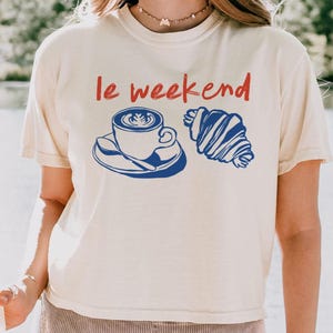 May include: Cream-colored t-shirt with the text "le weekend" in red. A blue graphic of a coffee cup and croissant is printed below the text. The shirt is paired with ribbed shorts.