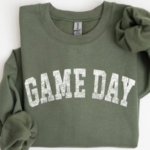May include: An olive green sweatshirt with the text "GAME DAY" in distressed white lettering. The crew neck sweatshirt is folded, with the sleeves rolled up. The Gildan brand tag is visible on the inside of the collar.