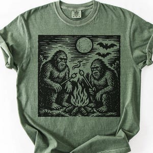 May include: Olive green t-shirt featuring a black graphic of two Bigfoot creatures roasting marshmallows over a campfire under a full moon and bats. The shirt has a crew neck and short sleeves.