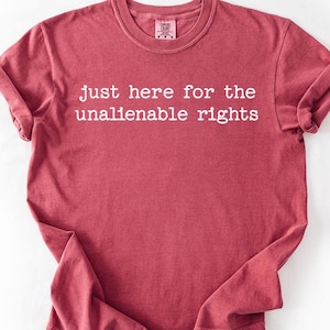 May include: A maroon-colored t-shirt with the text "just here for the unalienable rights" in white. The shirt has a crew neck and rolled-up sleeves. The shirt is made of a soft, comfortable material.