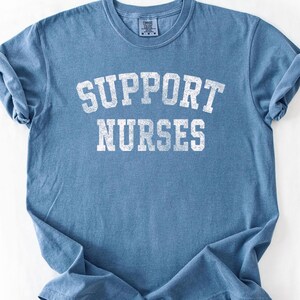 May include: A blue short-sleeved t-shirt with a crew neckline. The front features the text "SUPPORT NURSES" printed in a distressed white font. The fabric has a slightly faded, textured appearance with rolled sleeves and a standard fit.