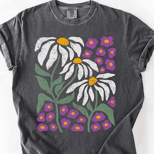 May include: Dark gray t-shirt featuring a floral design. The graphic includes large white daisies with yellow centers, green stems and leaves, and small purple flowers. The shirt has a crew neck and rolled-up sleeves.