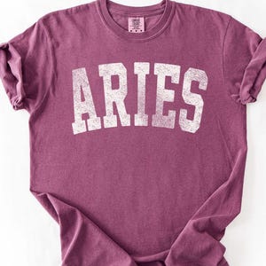 May include: A maroon t-shirt with the word "ARIES" printed in distressed white block letters. The shirt has short sleeves and a crew neck. The fabric is slightly wrinkled, giving it a casual look.