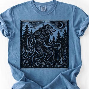 May include: A blue t-shirt featuring a black graphic of a werewolf in a forest under a crescent moon. The werewolf is depicted in a stylized, vintage illustration style. The shirt has a crew neck and short sleeves.