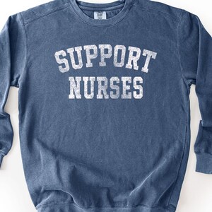 May include: A blue long-sleeve crewneck sweatshirt with a distressed white graphic print on the chest that reads SUPPORT NURSES in a bold, arched, block-style font. The garment features a ribbed collar, cuffs, and hem.
