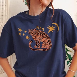 May include: Navy blue t-shirt featuring a stylized tiger illustration in orange and black, with the word "RAA" above the tiger. The design includes stars and a crescent moon. A casual, comfortable tee.